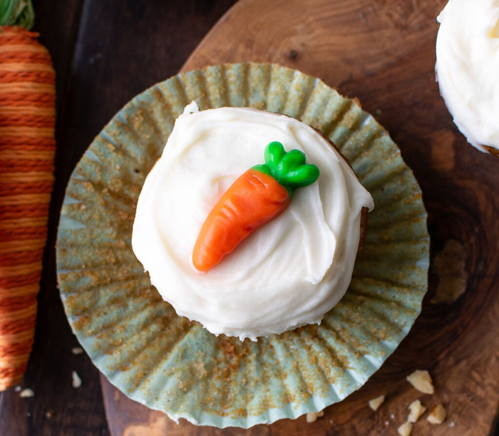 Close up of the top of a carrot cupcake with cream cheese frosting and a carrot shaped candy on top.