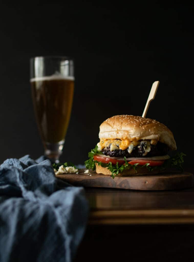 A Buffalo wing sauce burger on a cutting board draped with a blue linen napkin, and a glass of beer in the background.