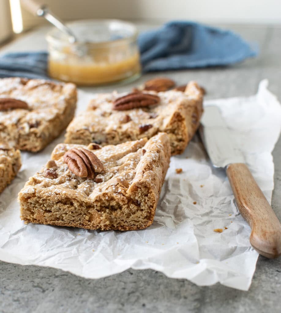 3 butter pecan bars on a slight of parchment with a wood handled knife beside them, and a jar of caramel sauce in the background.