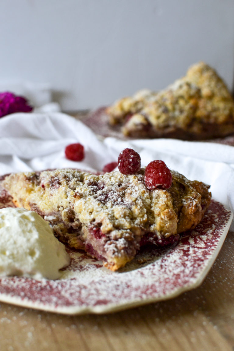 A white chocolate raspberry scone sprinkled with icing sugar and fresh raspberries with clotted cream beside