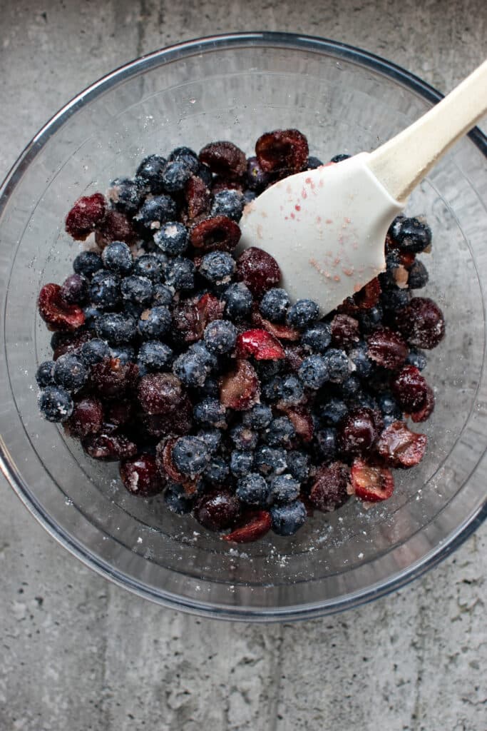 A bowl of blueberries and cherries combined with sugar, with a spatula in the bowl leaning against the edge.