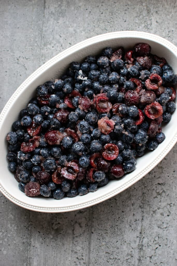 Blueberries and cherries combined with sugar in an oval ceramic baking dish.