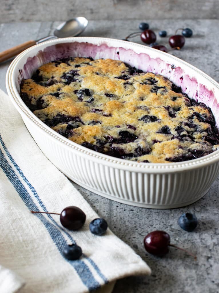 A freshly baked blueberry cherry cobbler in a oval shaped white baking dish.