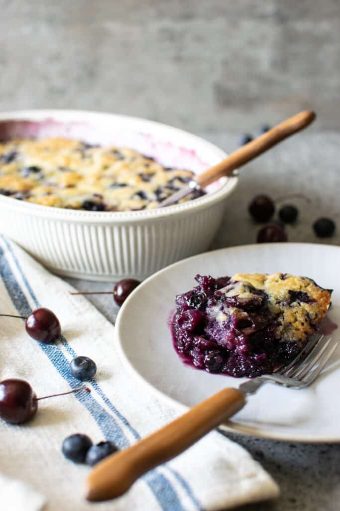 A plate of blueberry cobbler in the foreword with a baking dish with more cobbler is in the background.