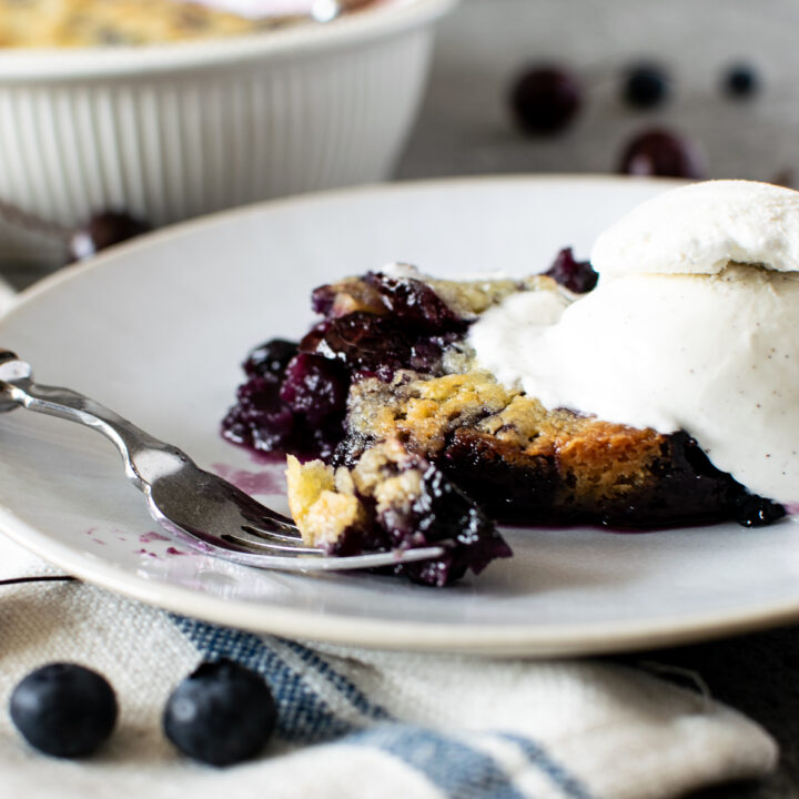 A fork with a piece of blueberry cherry crumble sits on a plate beside the full piece crumble with a scoop of ice cream on top.