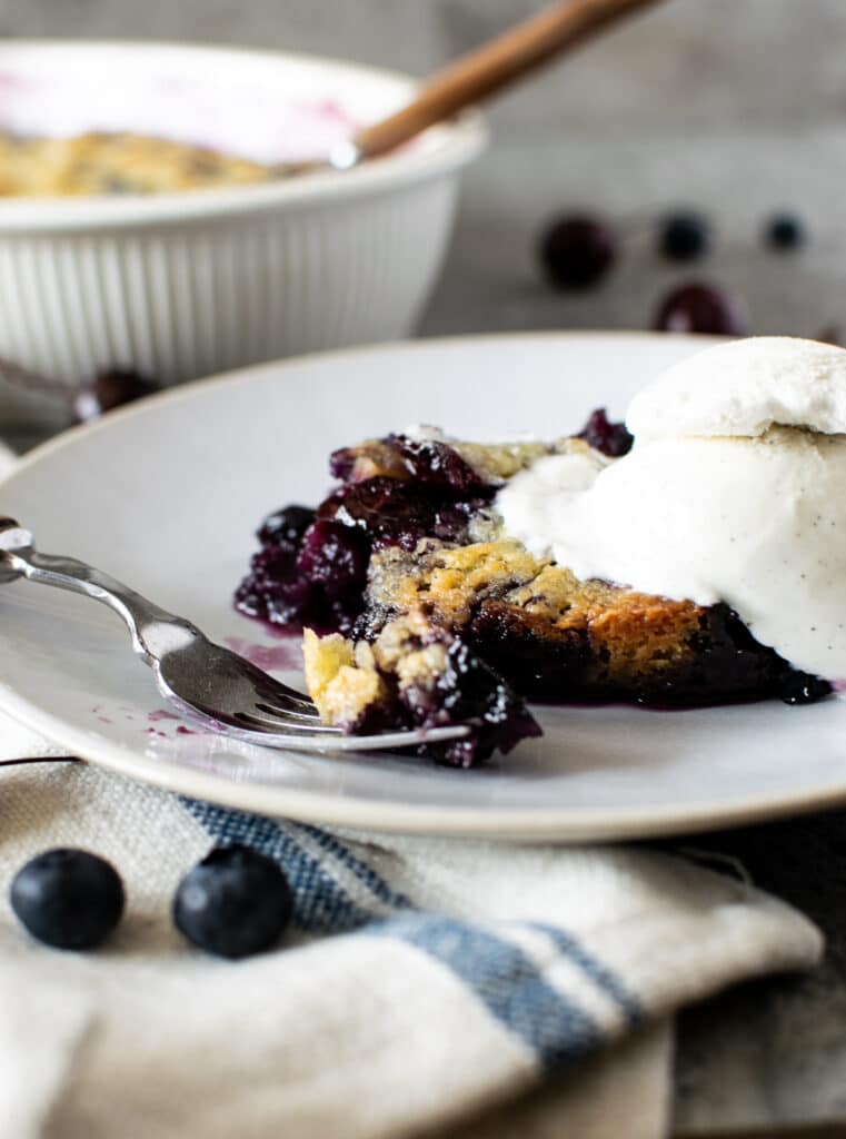 A fork with a piece of blueberry cherry crumble sits on a plate beside the full piece crumble with a scoop of ice cream on top.