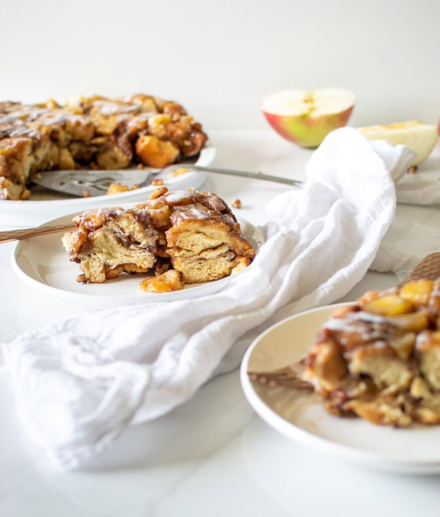 An apple cake in the background, with 2 individual slices in the foreground on white plates.