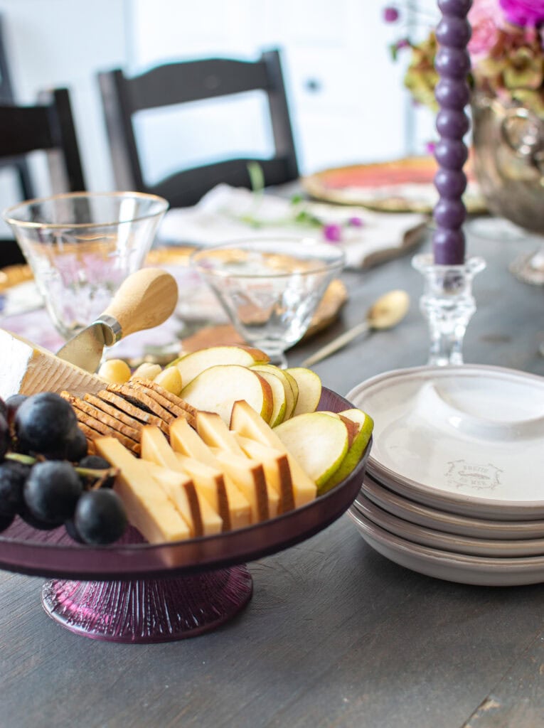 A small cheese platter on a purple cake plate, with crackers, cheese, purple grapes and sliced pears.