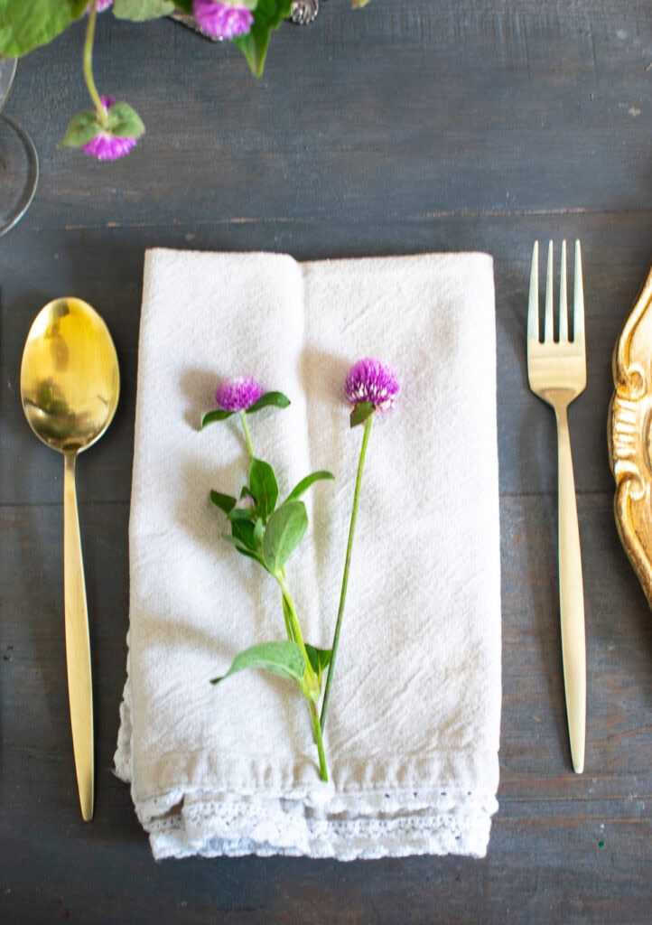 Close up of a cream coloured napkin with lace trim topped with two tiny purple flowers.  It is flanked by a gold spoon and fork.