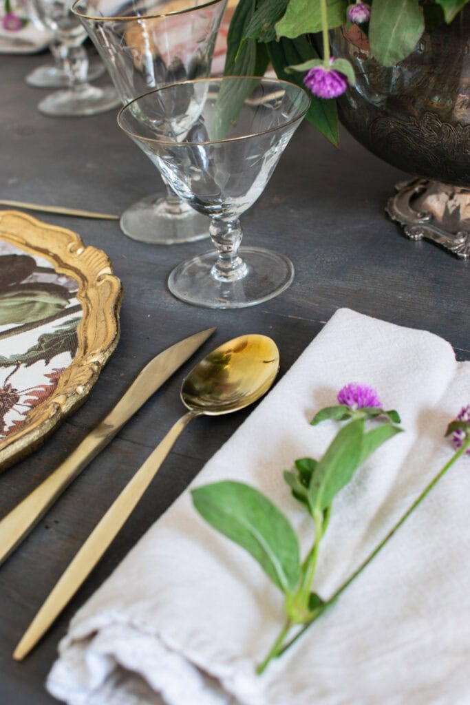 A close up photo of simple gold flatware beside a cream coloured napkin with little purple fresh flowers on top.