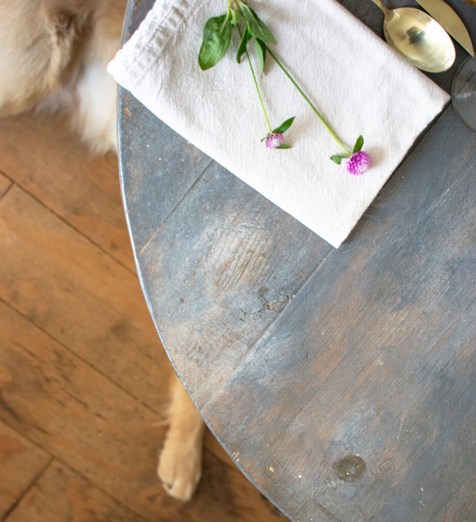 A photo of the edge of table with a napkin close to the edge, and a dog's paw sticking out from underneath the tables edge.