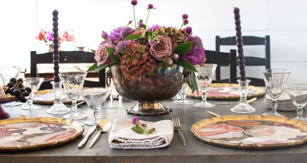 A dinner table set with a purple floral centrepiece, floral patterned charger plates and purple candles.