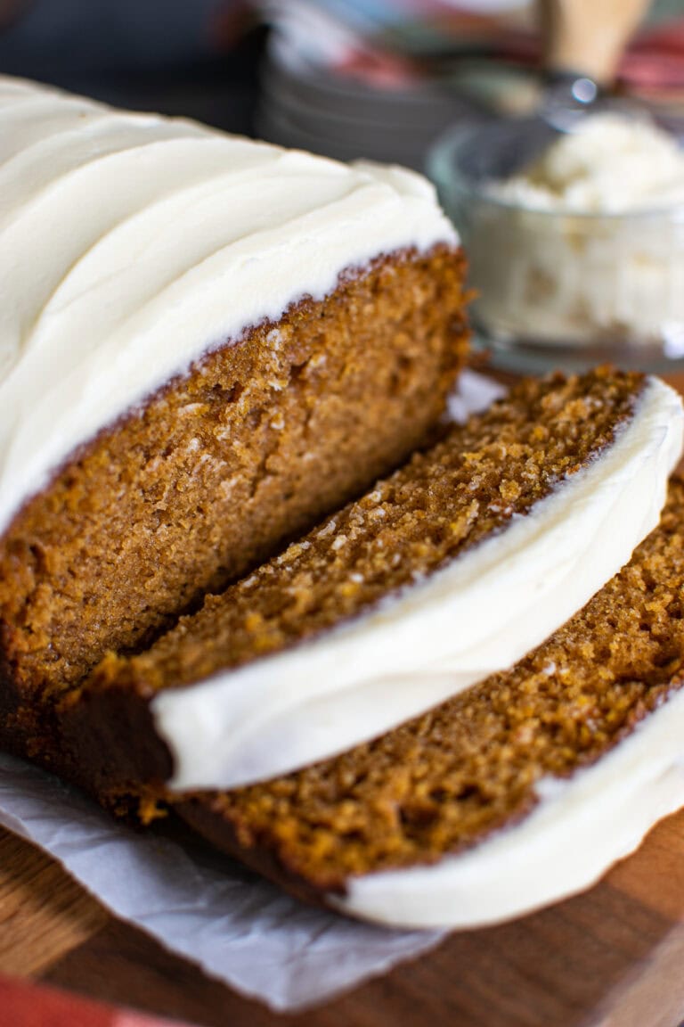 Pumpkin bread with cream cheese frosting with two slices cut and stacked in front of the loaf.