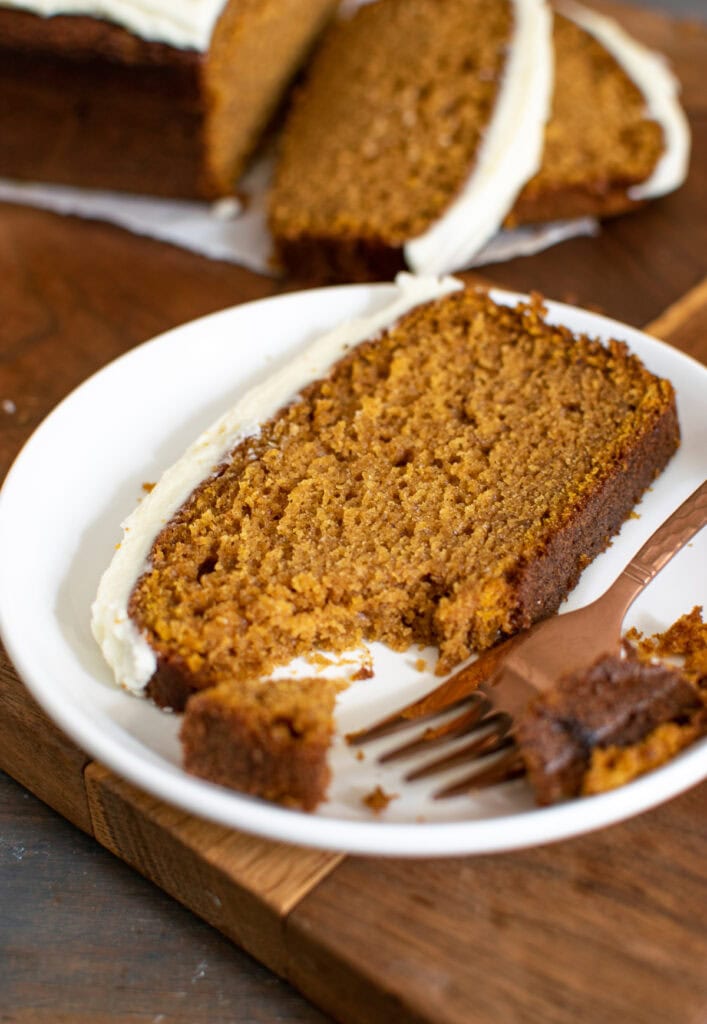 A slice of pumpkin bread with cream cheese frosting on a plate, with a copper fork.