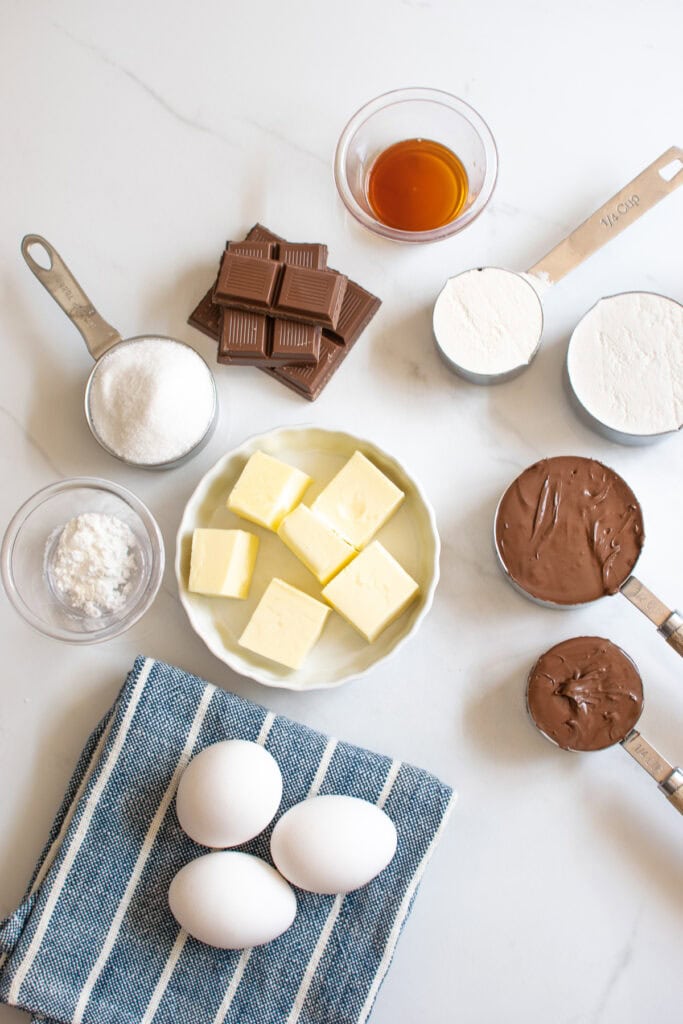 Overhead view of ingredients to make chocolate hazelnut cake including butter, chocolate, hazelnut spread, flour and eggs, on top of a blue and white dish towel.