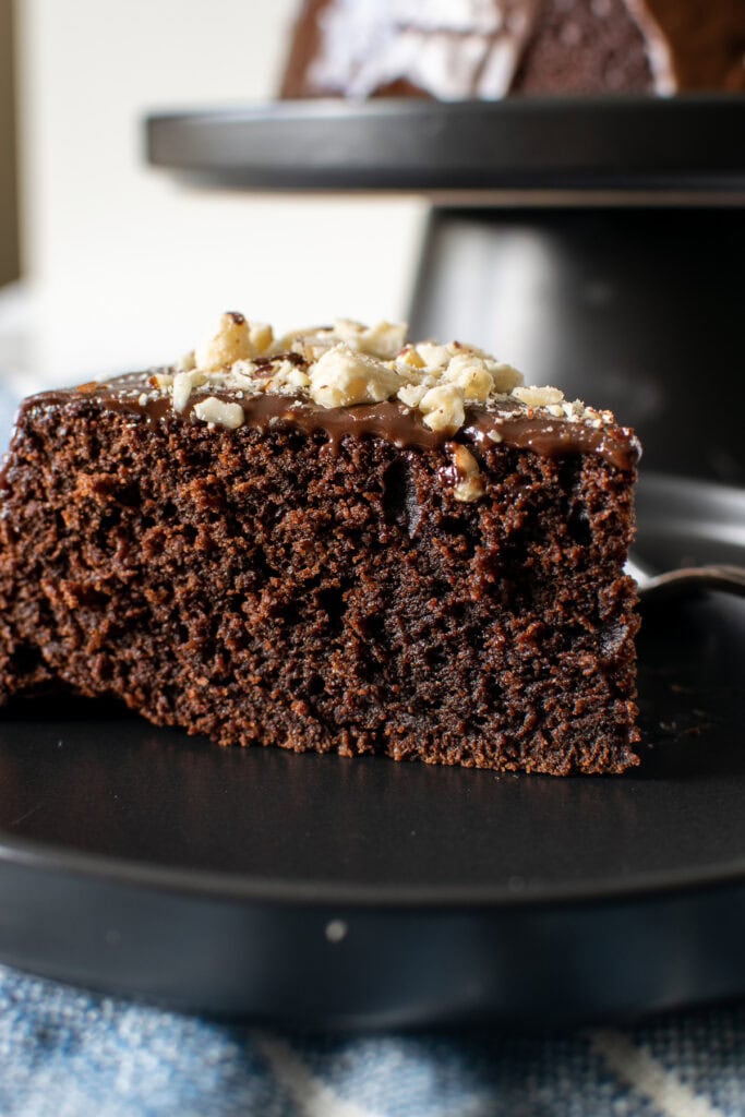 A close up photo of chocolate hazelnut cake with ganache frosting on a black plate.