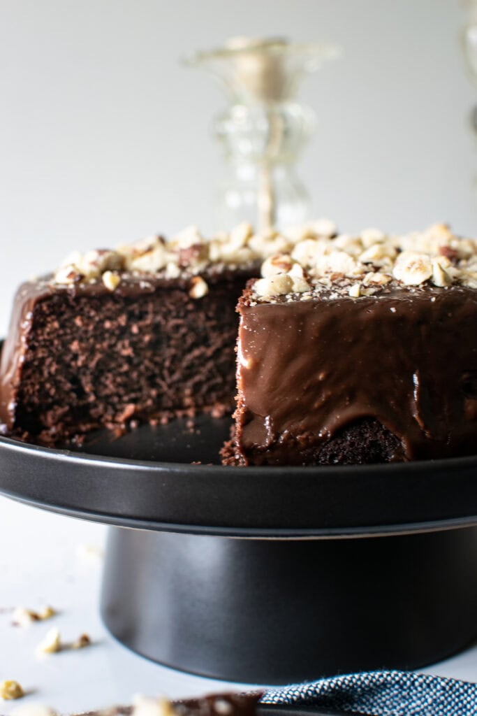 A chocolate cake with hazelnuts on top of ganache frosting , and one slice cut out, sits on a black cake stand.