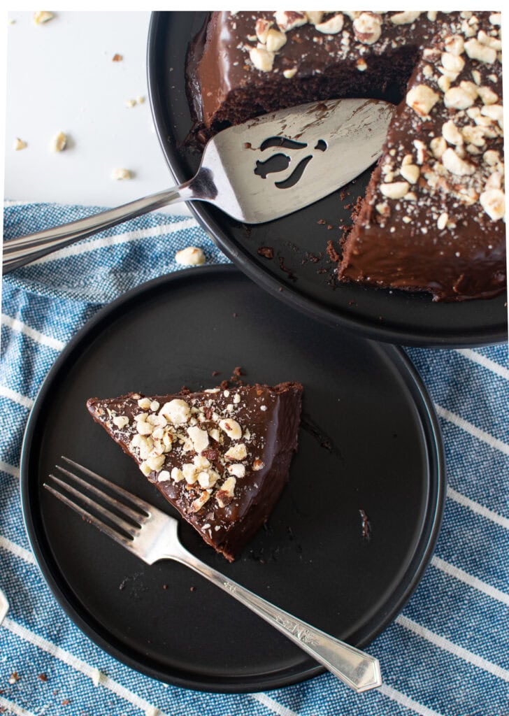 Overhead view of a slice of chocolate cake topped with hazelnuts on a black plate with a silver fork beside it.