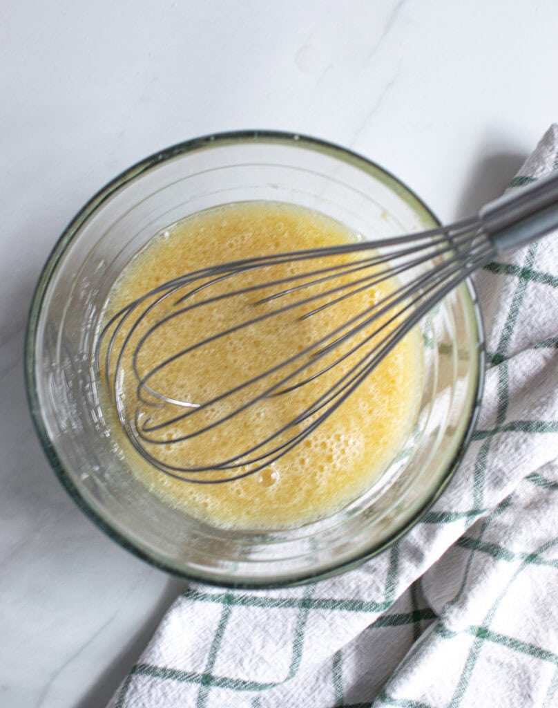 Egg, maple syrup, milk, vanilla and oil that has been whisked together in a glass bowl, with the whisk still in the bow.