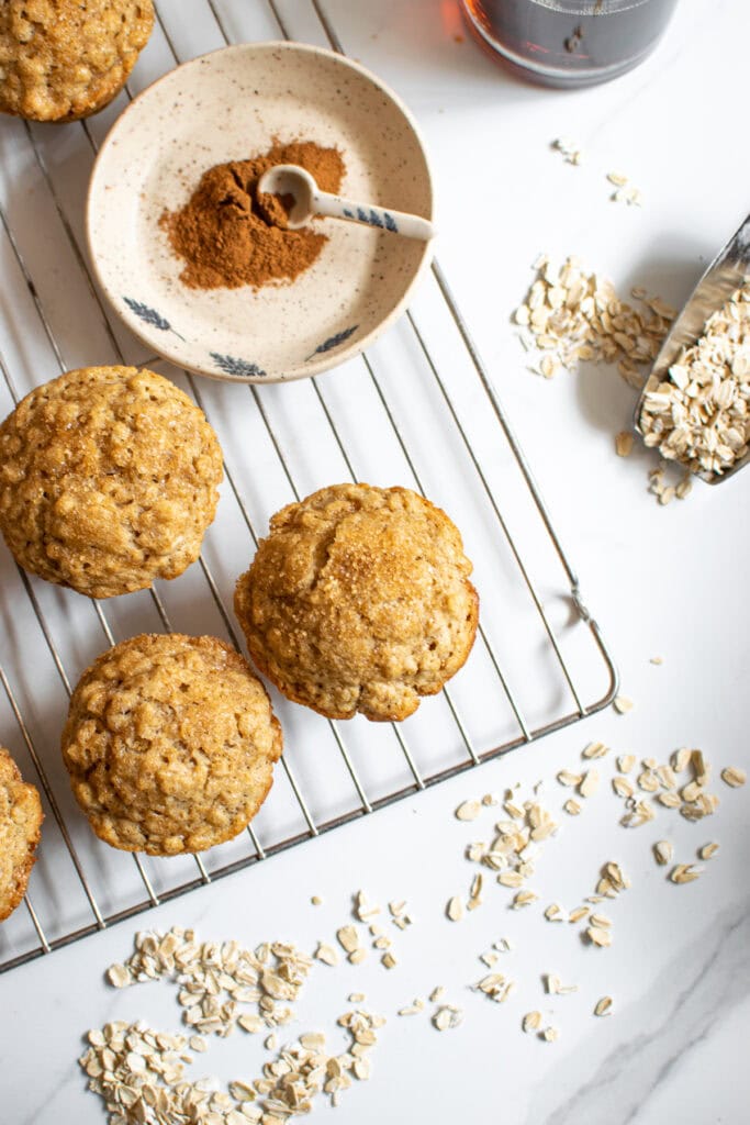 Overhead photo of 3 muffins on a cooling rack with a little plate with cinnamon on it and a scoop full of oats.