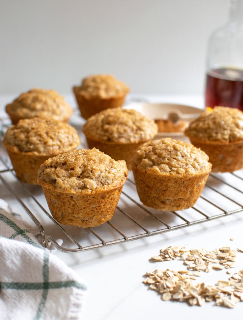 7 Oatmeal Cinnamon Muffins on a cooling rack, with oats sprinkled on the counter and a bottle of maple syrup in the background.