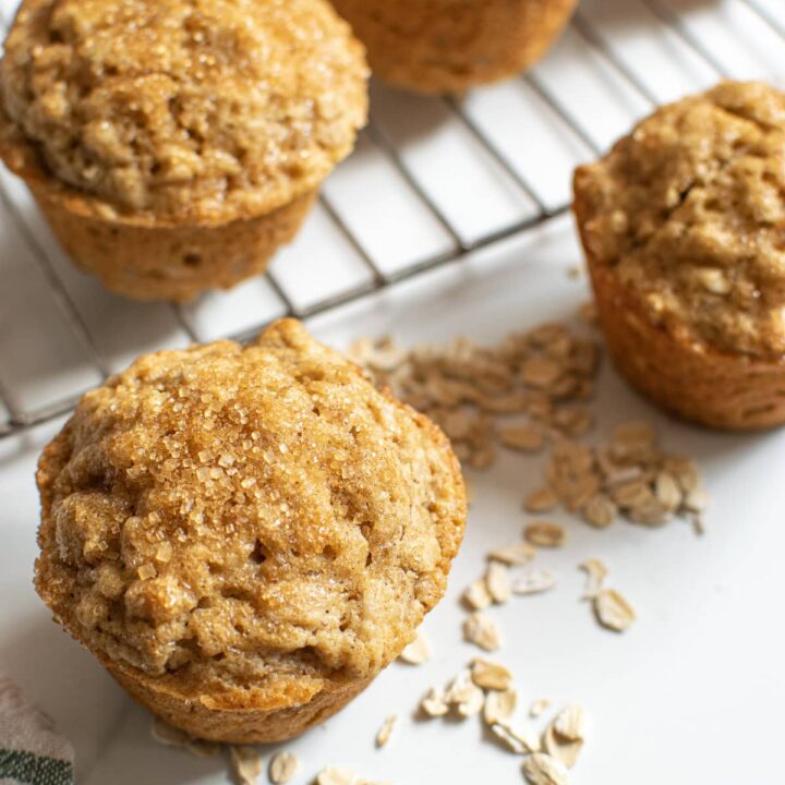 A cooling rack with two muffins on it, plus two muffins in front of it, with oats sprinkled around them.