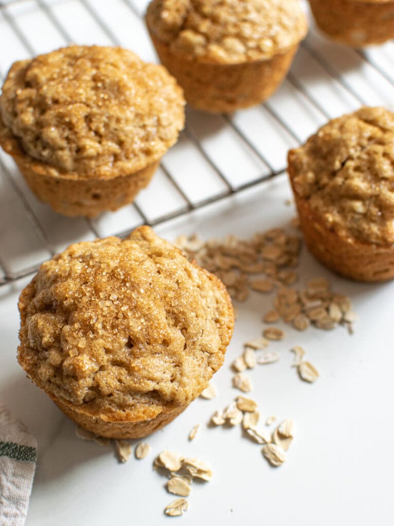 A cooling rack with two muffins on it, plus two muffins in front of it, with oats sprinkled around them.