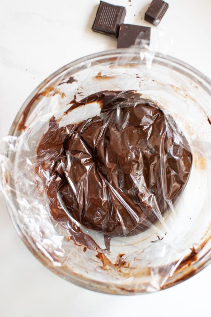 Chocolate ganache with plastic film over top of it in a mixing bowl to prevent a skin from forming.