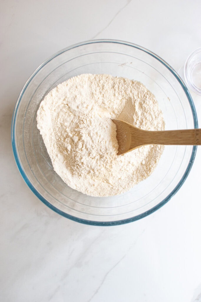 An overhead view of the dry ingredients to make banana bread mixed together in a glass bowl with a wooden spoon