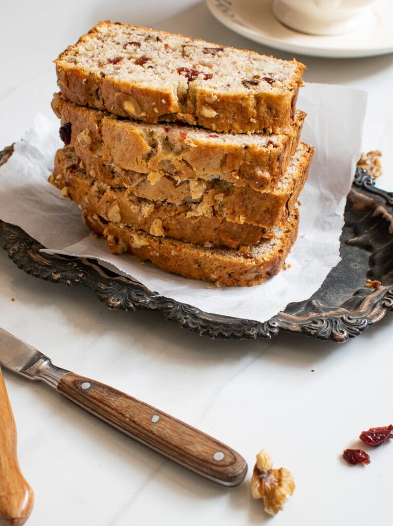 A stack of banana bread slices with cranberries and walnuts on a tarnished silver plate.