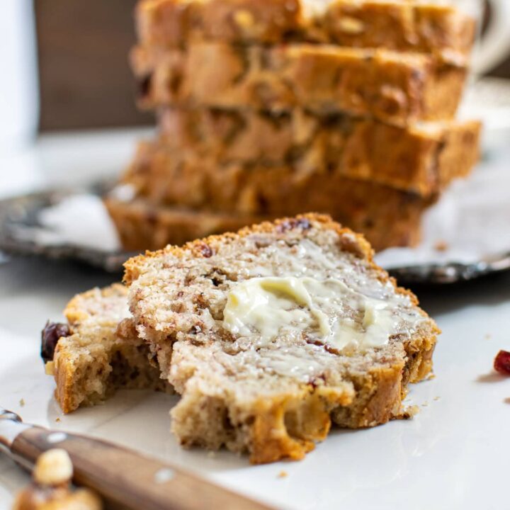 A slice of buttered banana bread with walnuts and cranberries in front of a stack of more slices.