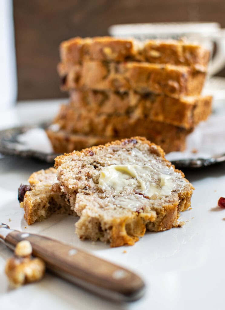 A slice of buttered banana bread with walnuts and cranberries in front of a stack of more slices.