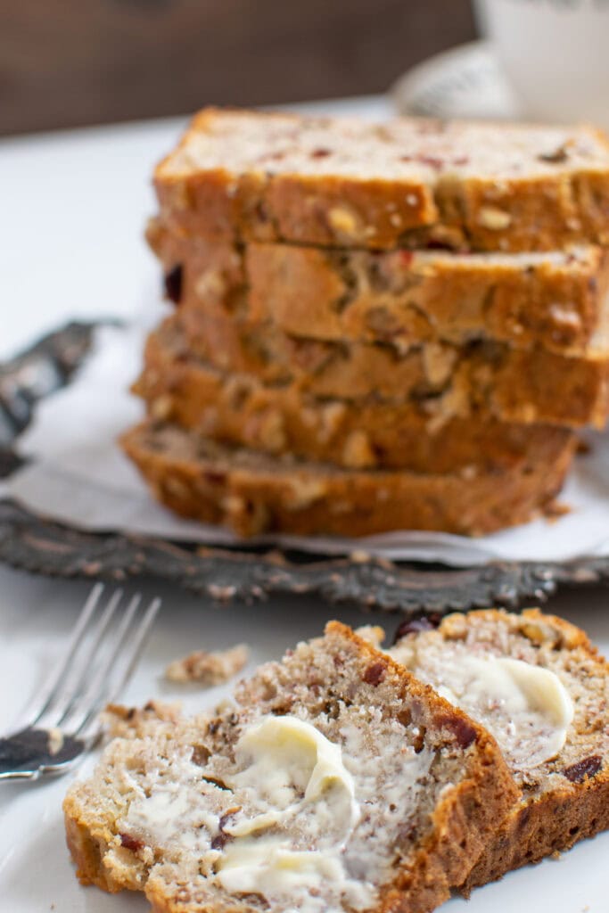 A stack of sliced cranberry banana nut bread with a buttered slice in front.