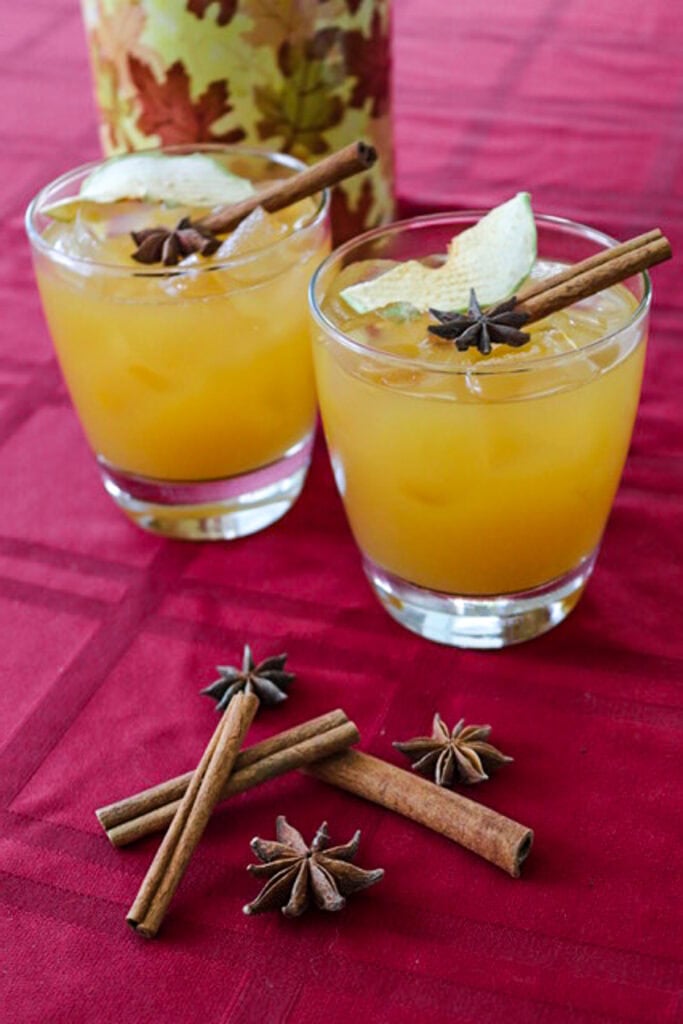 2 glasses of apple cider rum cocktail topped with cinnamon sticks and dried apple slices.