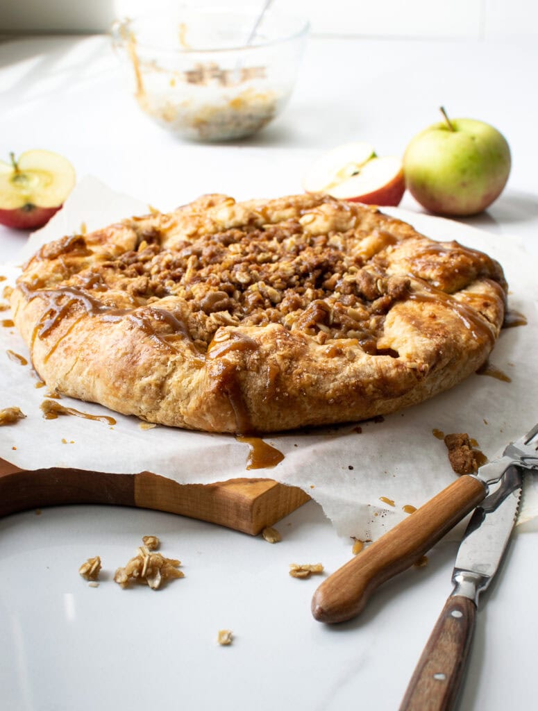 An apple and pear galette on a sheet of parchment with a knife and fork in the foreground and apples in the background.