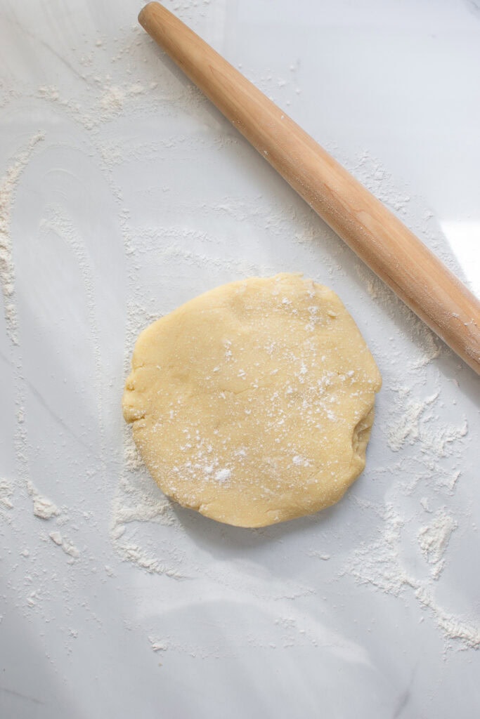 A disc of chilled sweet pastry dough on a floured counter and a rolling pin beside it.