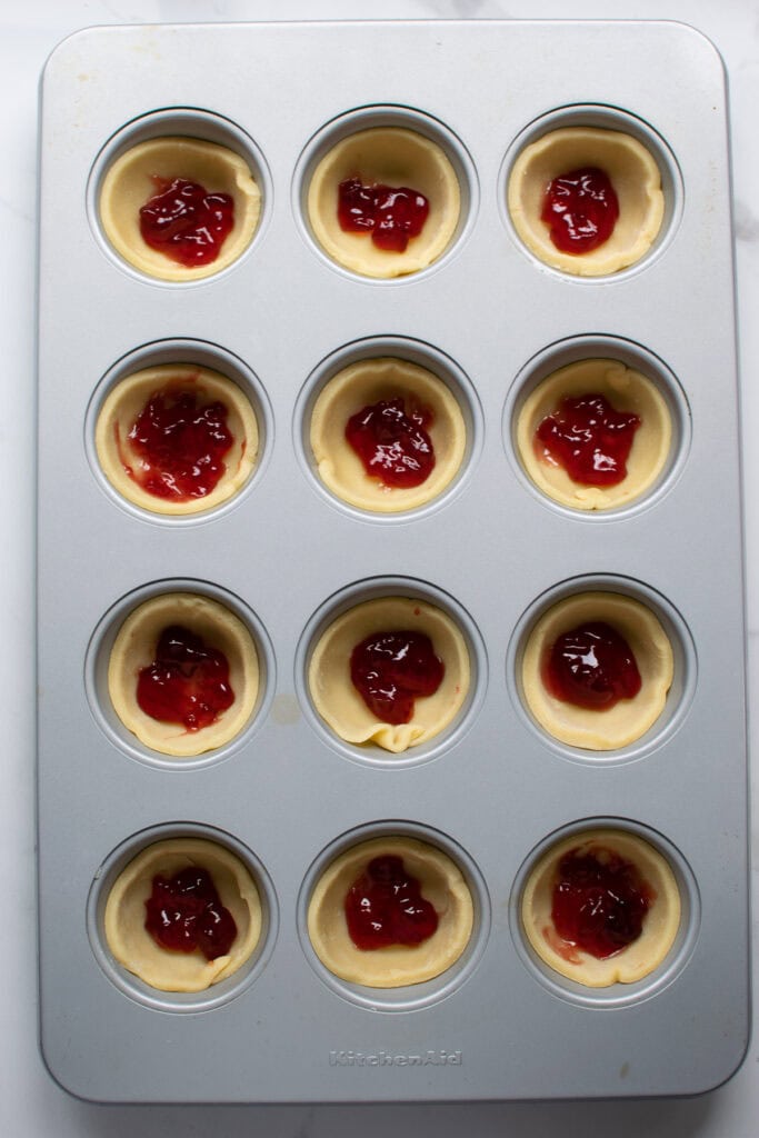 Overhead view of sweet pastry in muffin tins with dots of cherry jam in each one.