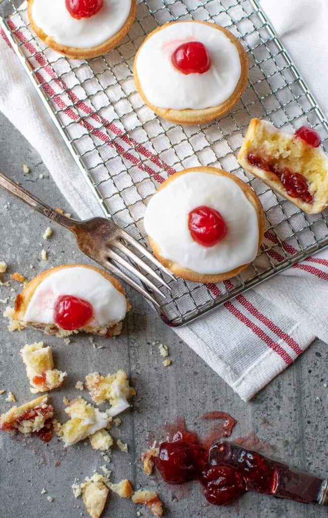 Overhead view of a cherry bakewell tart on a cooling rack, with one on the counter that is half eaten.