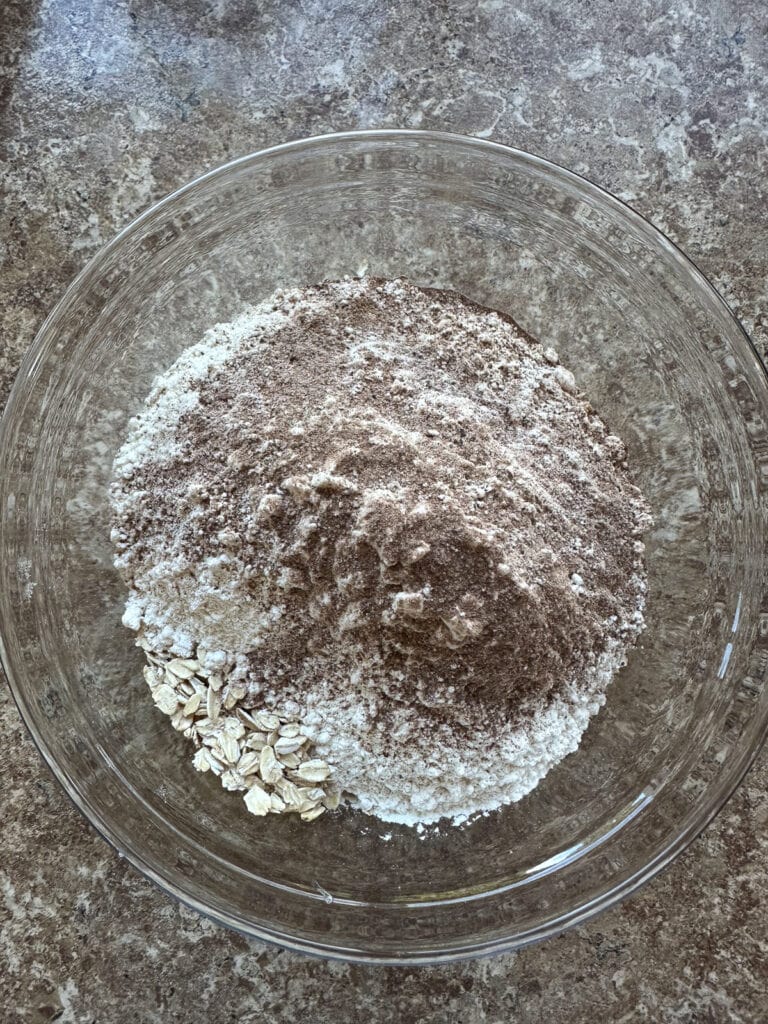 Layers of oats, flour and spices waiting to be stirred together in a glass mixing bowl.