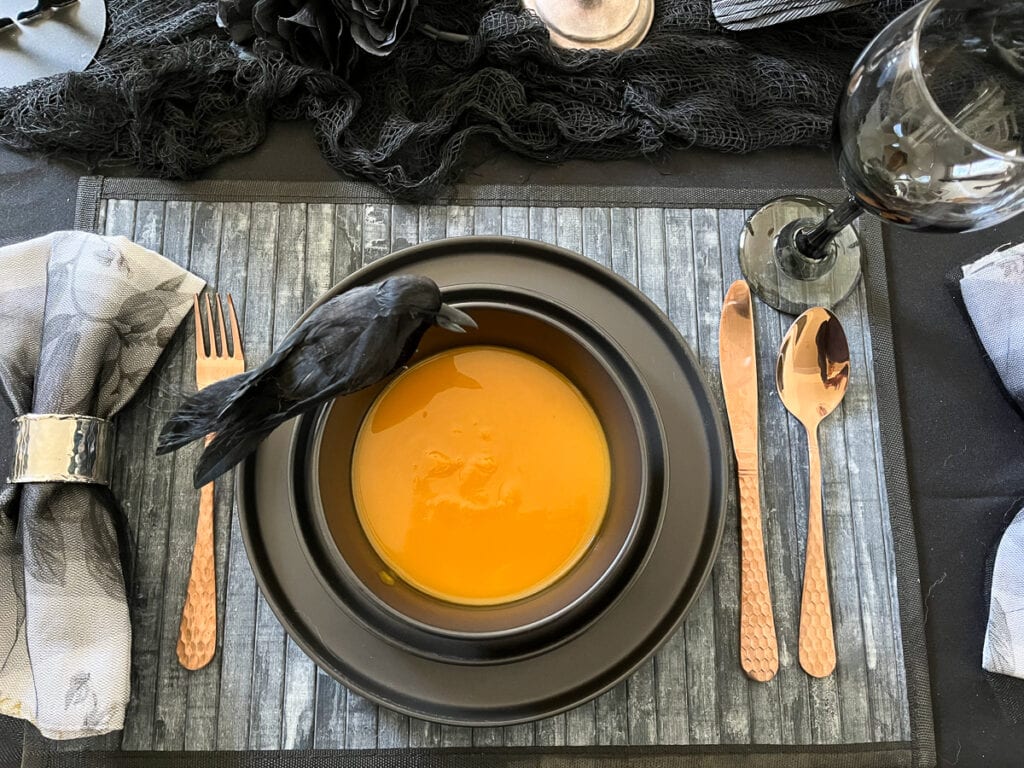 An overhead view of butternut squash soup served in black round bowls with a faux crow perched on the edge.