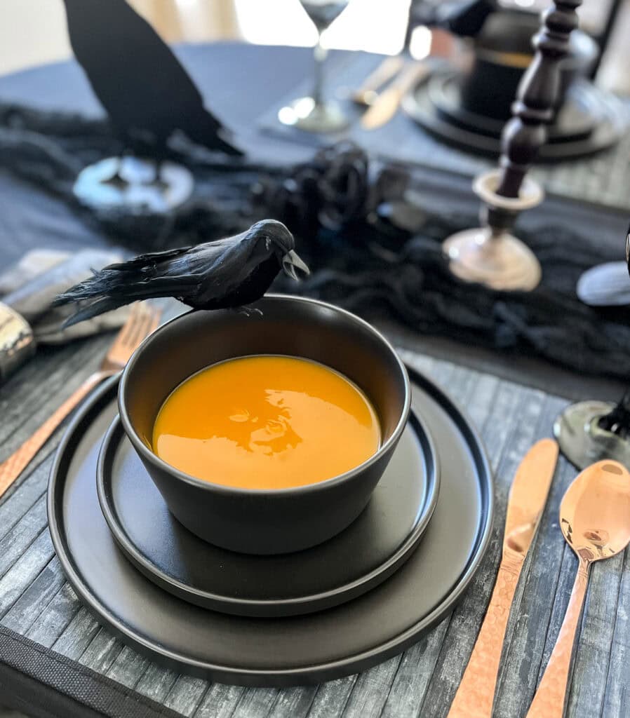 A black stoneware dinner plate stack with a bowl on top filled with butternut squash soup, with copper flatware beside it.