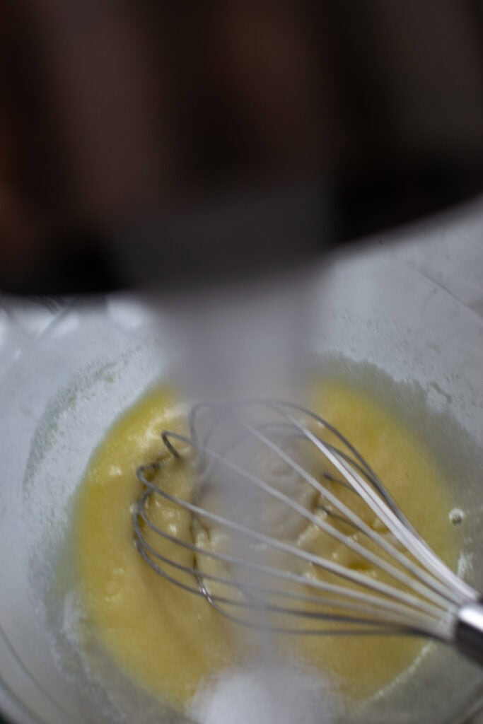 Sugar slowing being pouring into a bowl while whisking an egg.