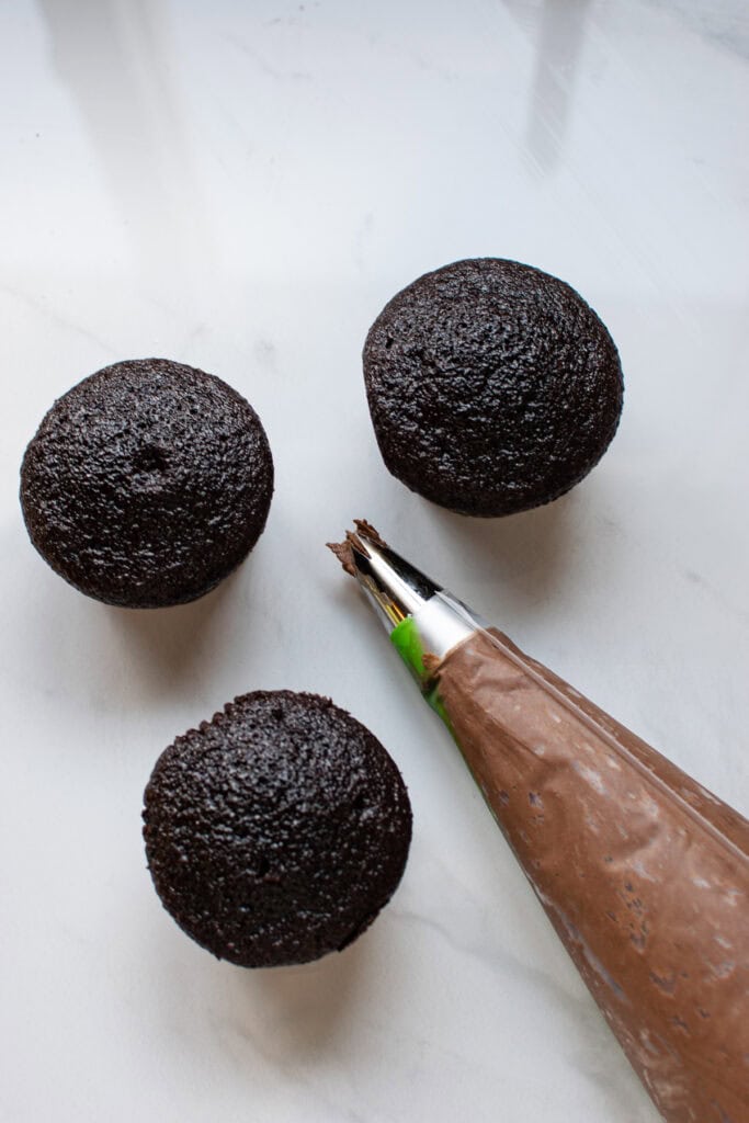 A piping bag filled with chocolate frosting with 3 cupcakes beside it.