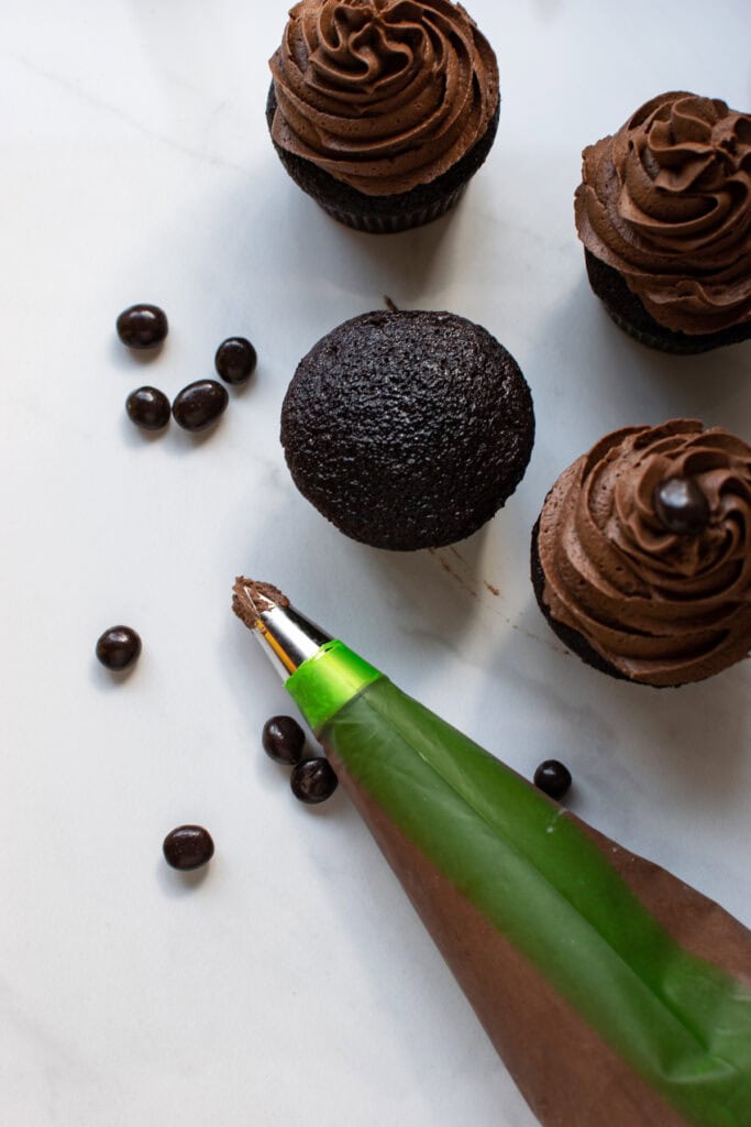 Chocolate frosting in a piping bag with 2 frosted cupcakes, 1 unfrosted and a sprinkle of chocolate covered coffee beans.