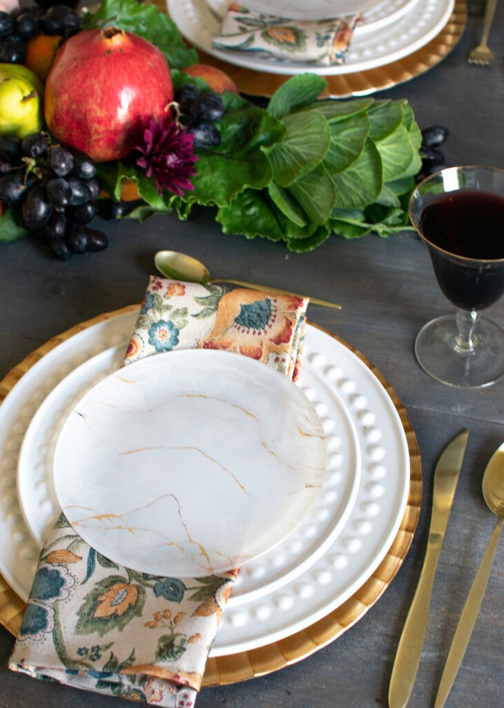 A place setting with gold charger plate, white dinner and salad plates, and a gold marbled dessert plate. A floral napkin in fall colours sits under the dessert plate and gold flatware are on each side.