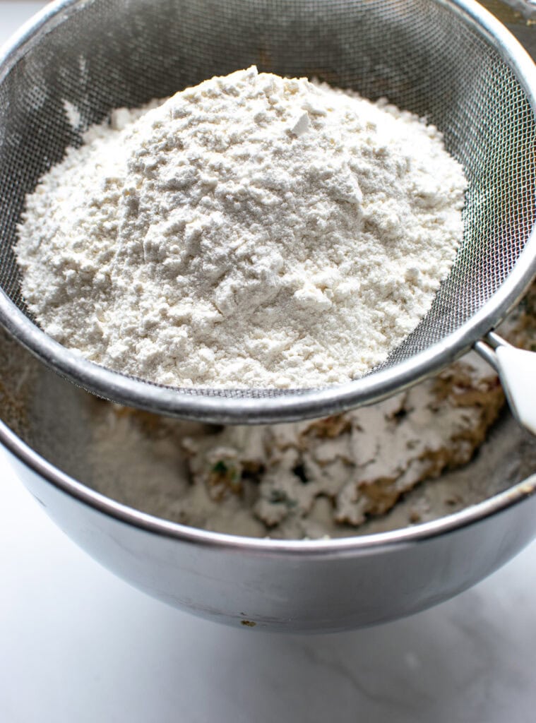 A sifter set over a mixing bowl and filled with flour and baking powder to be sifted into cookie dough.