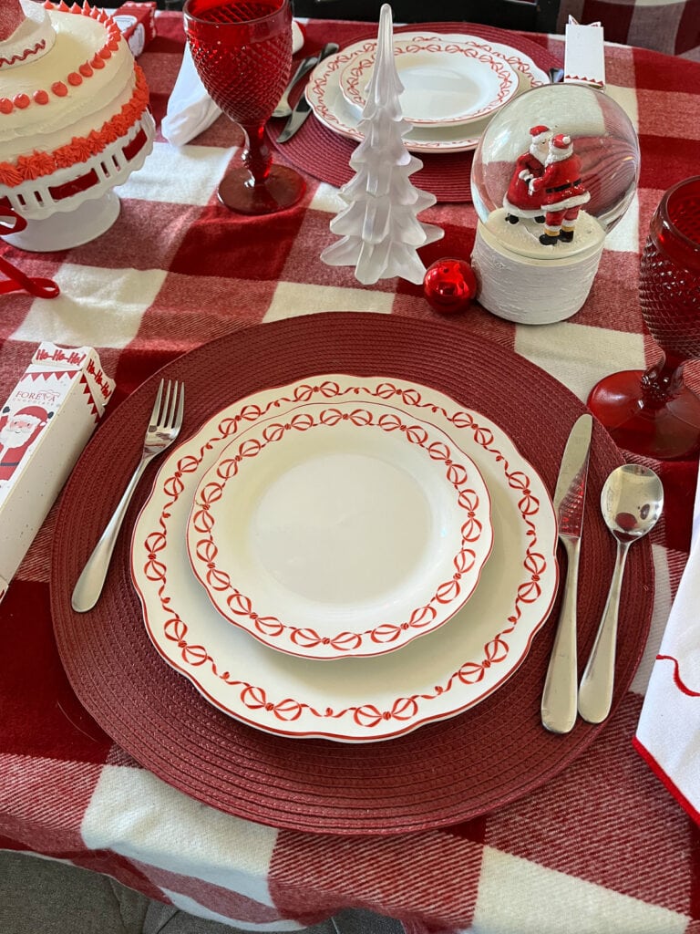 A red and white Christmas place setting on a dining table. Red and white check table cloth, round red placemat with white plates that have red bows around the edges.