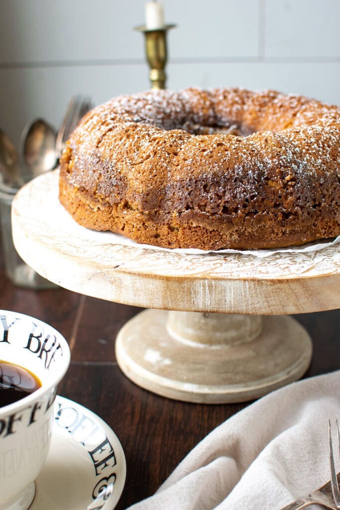 A coffee cake made in a  bundt pan sitting on a cake stand beside a cup of coffee.