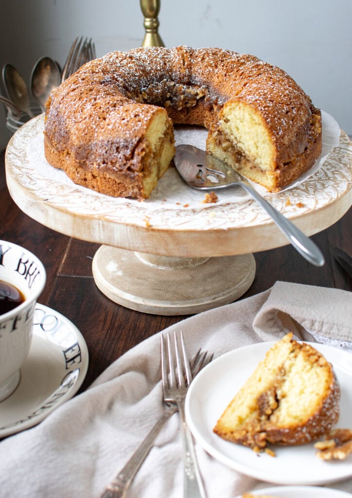 A cinnamon walnut coffee cake on a wood cake stand with one slice on a plate.