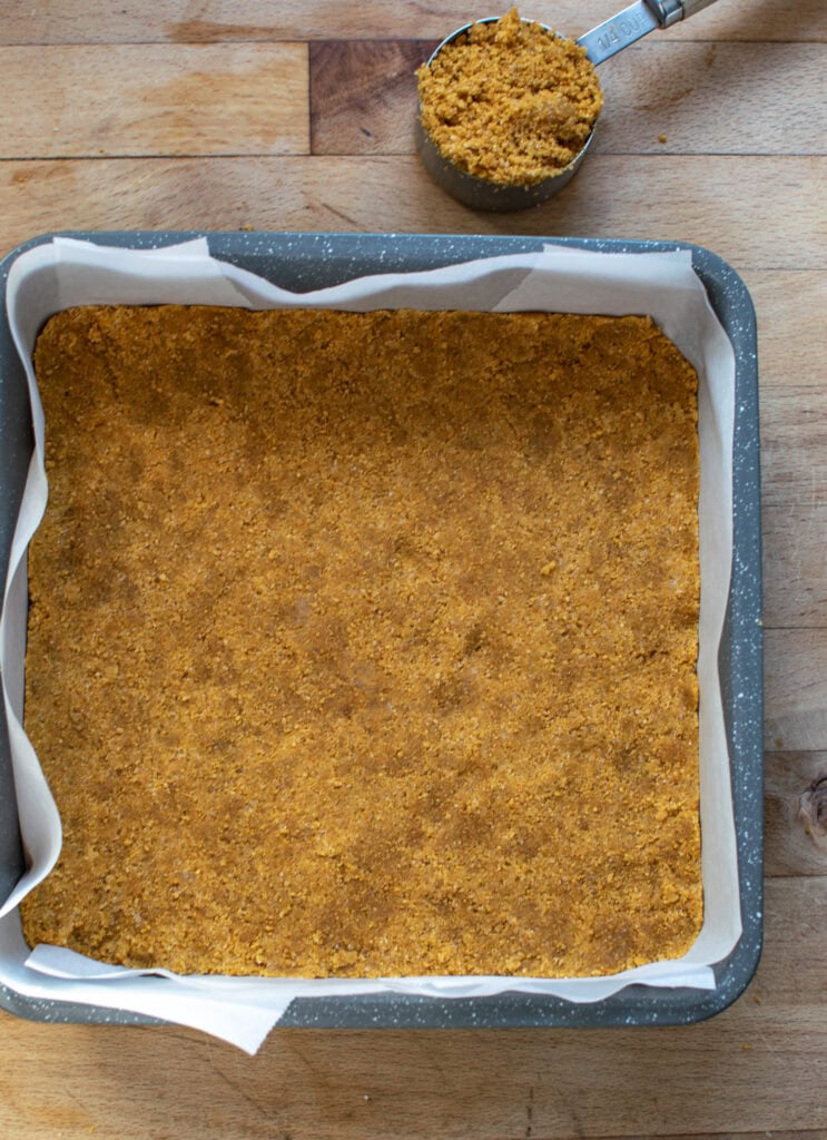 A nine inch square baking pan lined with parchment, with a graham cracker crust pressed into the pan.